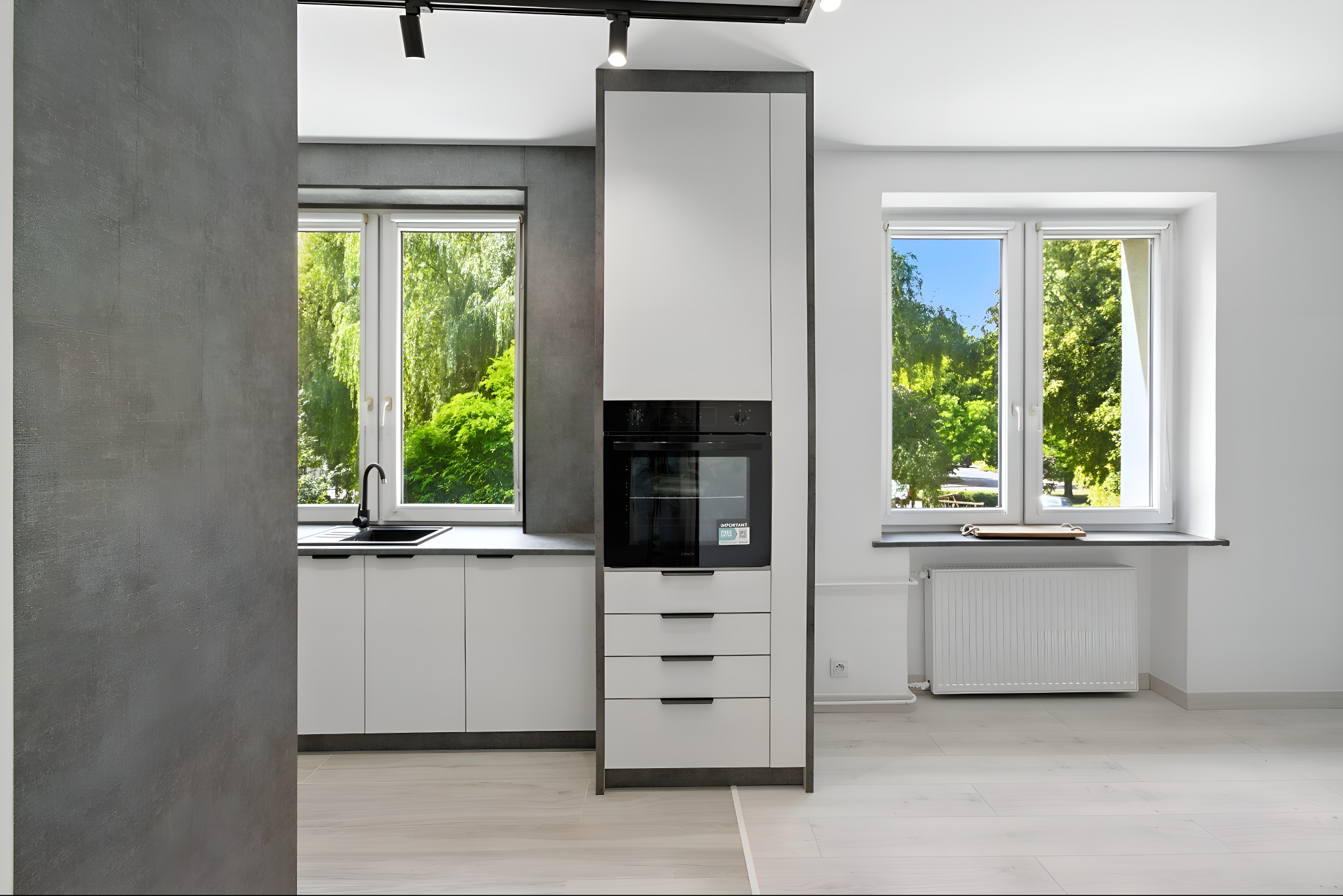 Jasna kuchnia z oknami i widokiem na zieleń. Minimalistyczne szafki, czarny zlew i piekarnik w zabudowie. Szare ściany i drewniana podłoga. Nowoczesny design.