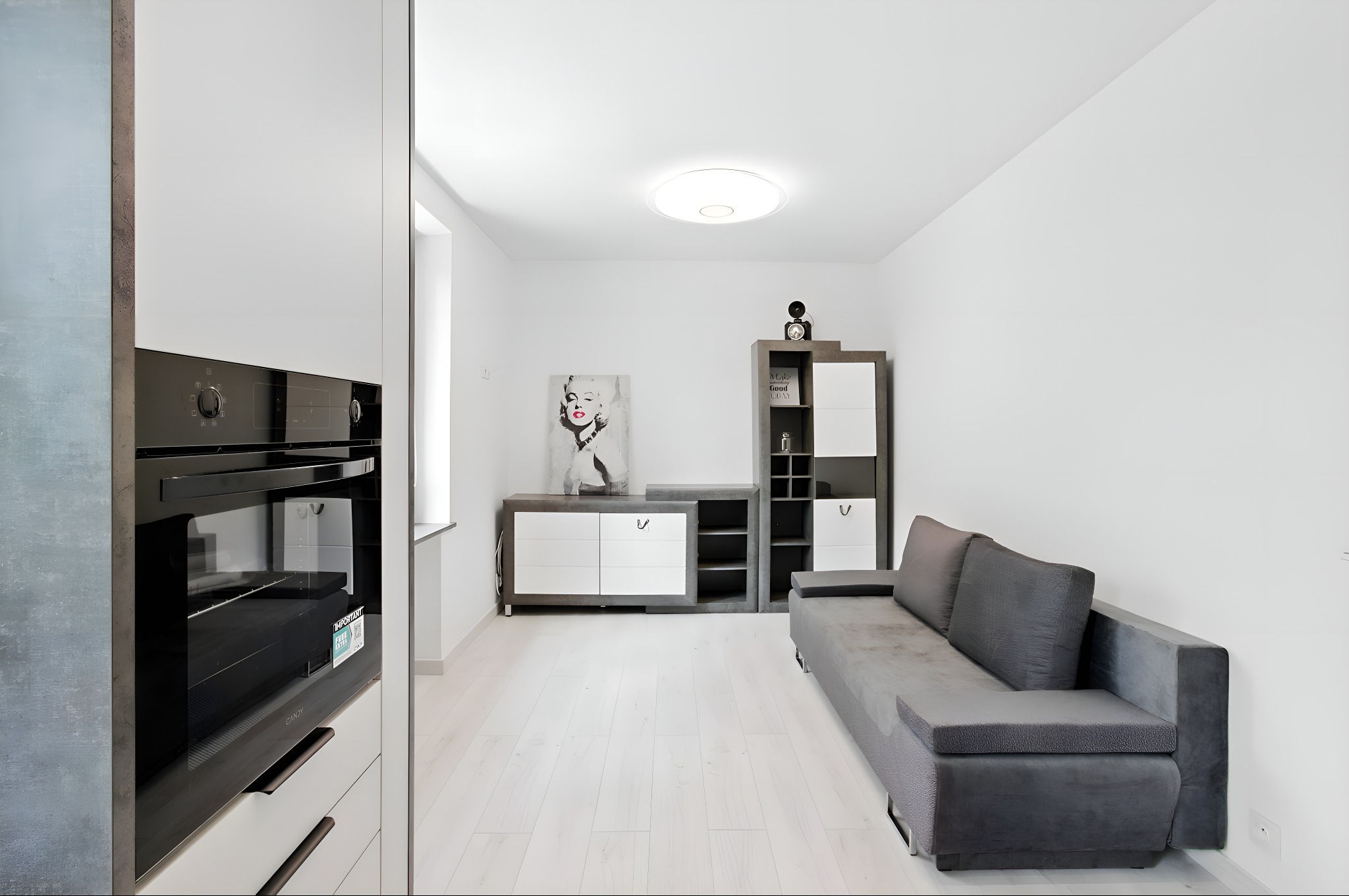 Jasne wnętrze salonu z szarą sofą i białymi meblami. Na ścianie obraz z Marilyn Monroe. Widoczny fragment zabudowy kuchennej z piekarnikiem.