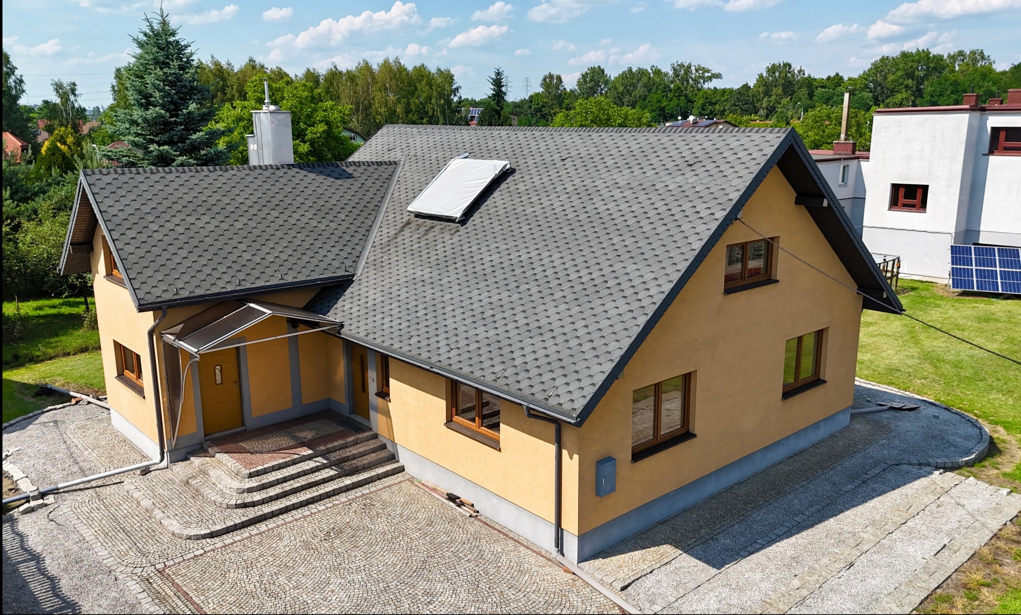 Żółty dom jednorodzinny z szarym dachem z gontów bitumicznych, widok z góry na brukowany podjazd i zielony ogród z panelami słonecznymi w tle.