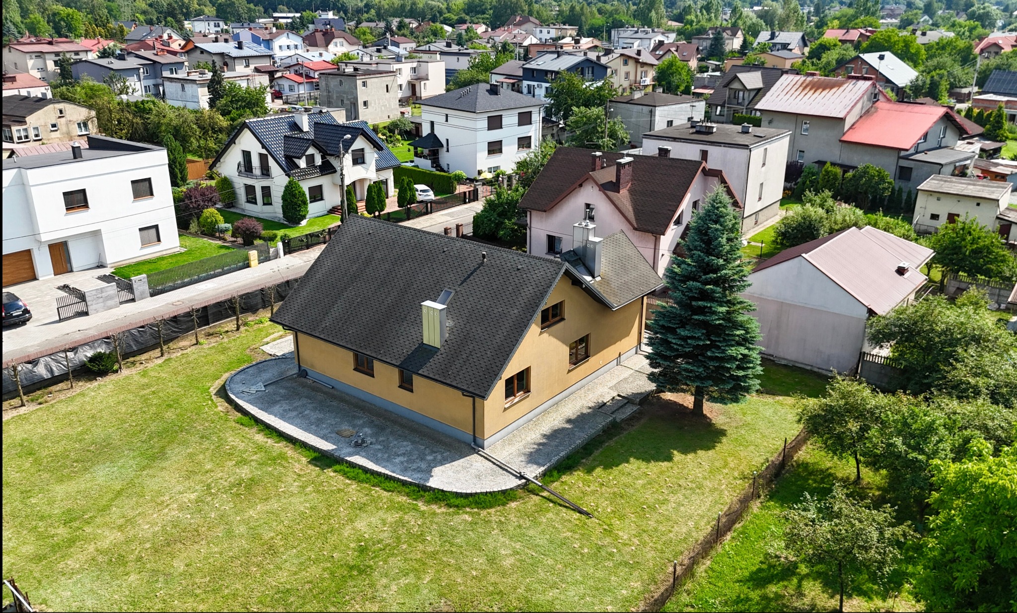 Żółty dom jednorodzinny z ciemnym dachem na tle zabudowy miejskiej, widziany z góry. Wokół domu trawnik i drzewo iglaste. Uwagę przykuwa wykończenie wokół budynku.