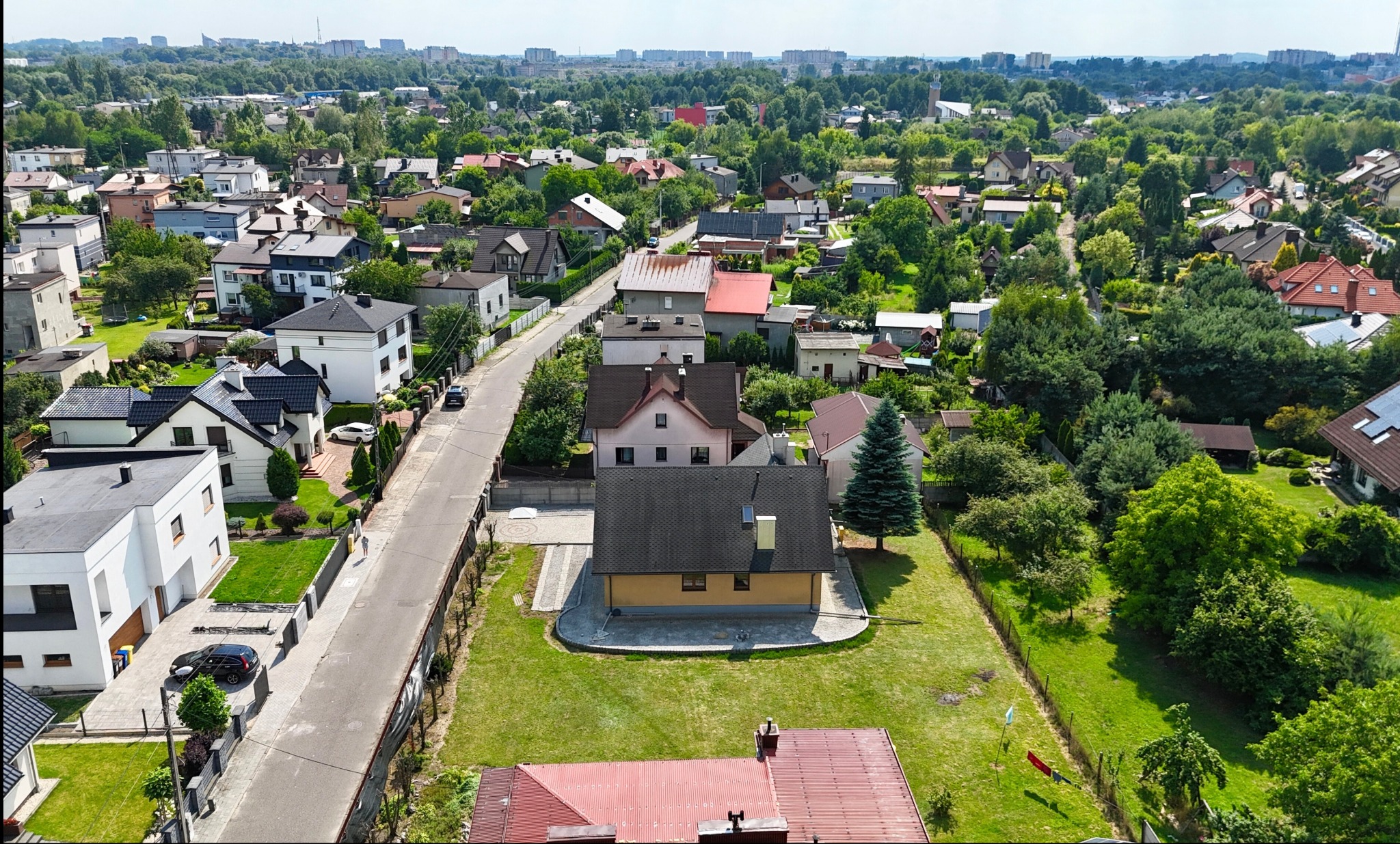 Panorama osiedla domów jednorodzinnych z lotu ptaka, zieleń drzew, zabudowa miejska w tle, słoneczny dzień, widok na ulicę i zadbane ogrody.