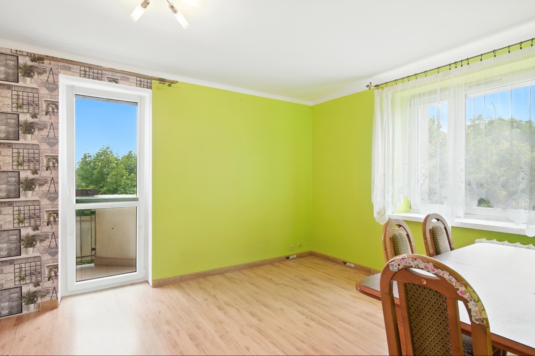 Jasnozielony pokój z balkonem i oknem z widokiem na zieleń. Parkiet na podłodze, stół z krzesłami. Tapeta w cegiełki na ścianie. Słoneczny dzień.