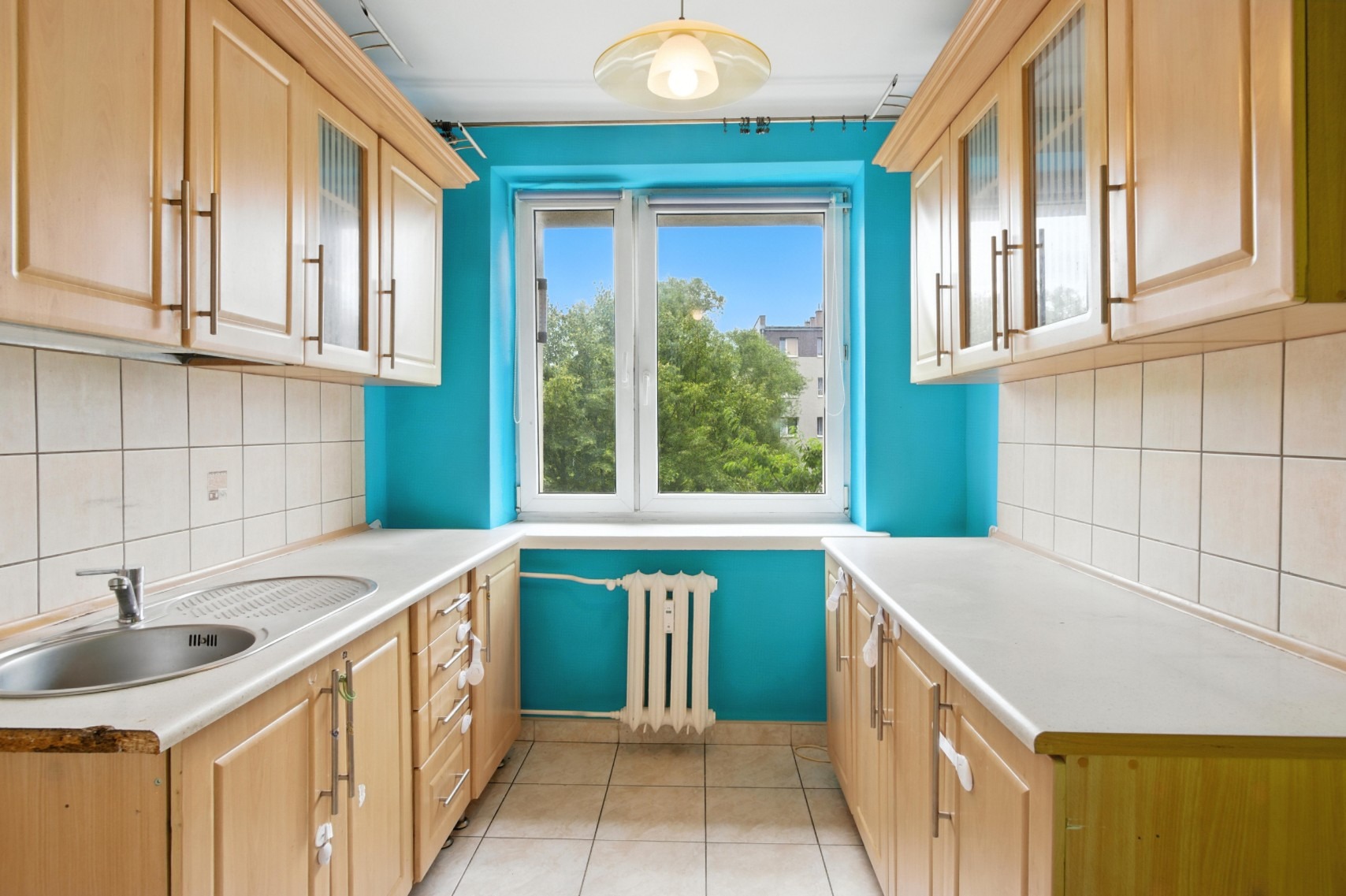 Jasna kuchnia z błękitną ścianą i widokiem na zieleń za oknem. Umeblowanie z drewnianymi szafkami, białe blaty i płytki. Centralnie umieszczone okno z grzejnikiem.