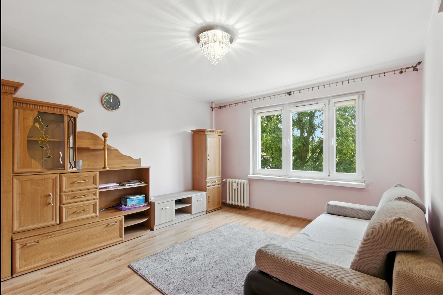 Jasny pokój z drewnianymi meblami, szarą sofą i oknem z widokiem na zieleń. Kryształowy żyrandol dodaje elegancji. Parkiet na podłodze.