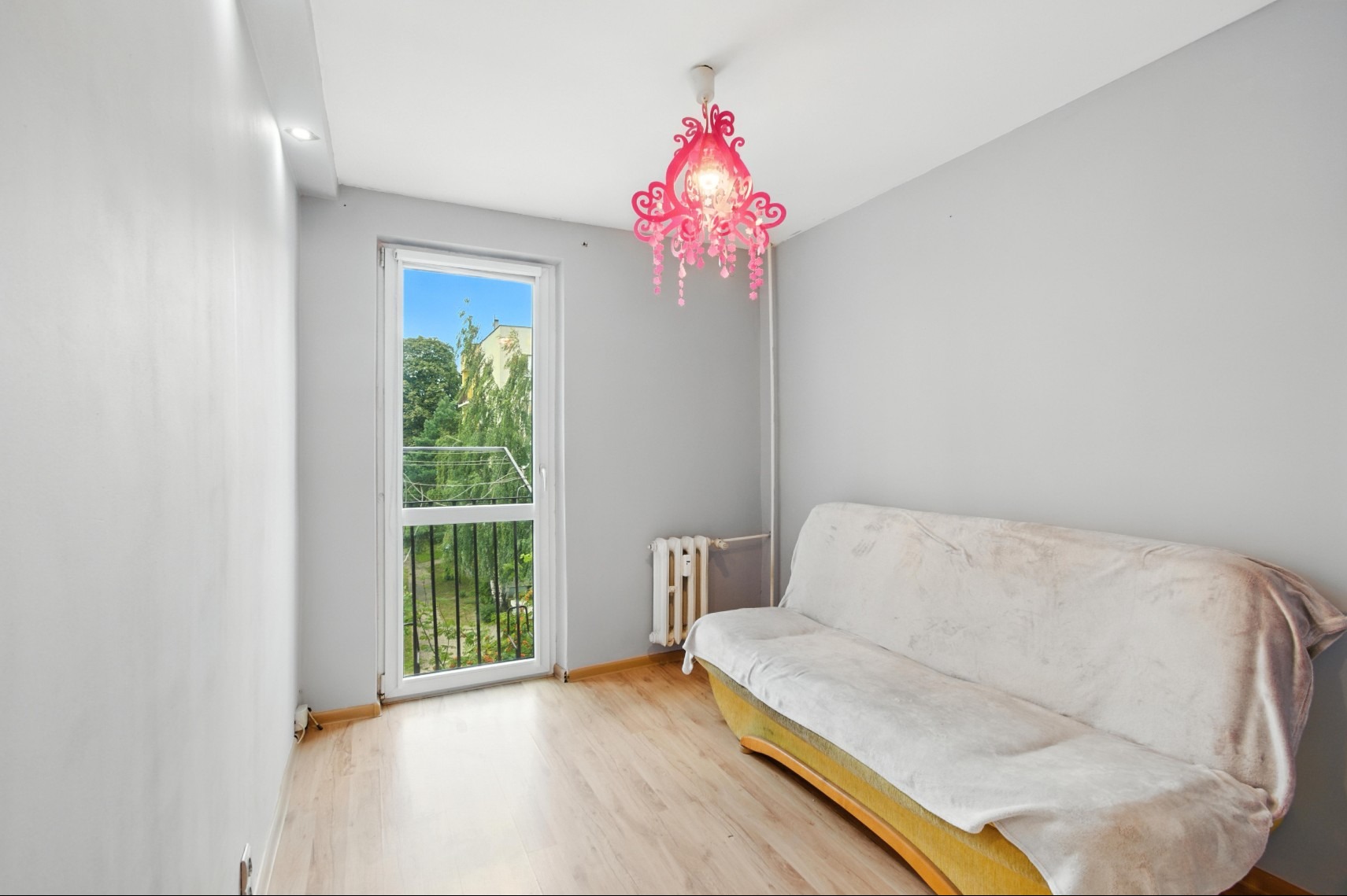 Jasne wnętrze pokoju z balkonem i parkietem, sofa przykryta narzutą, różowy żyrandol. Widok na zieleń za oknem.