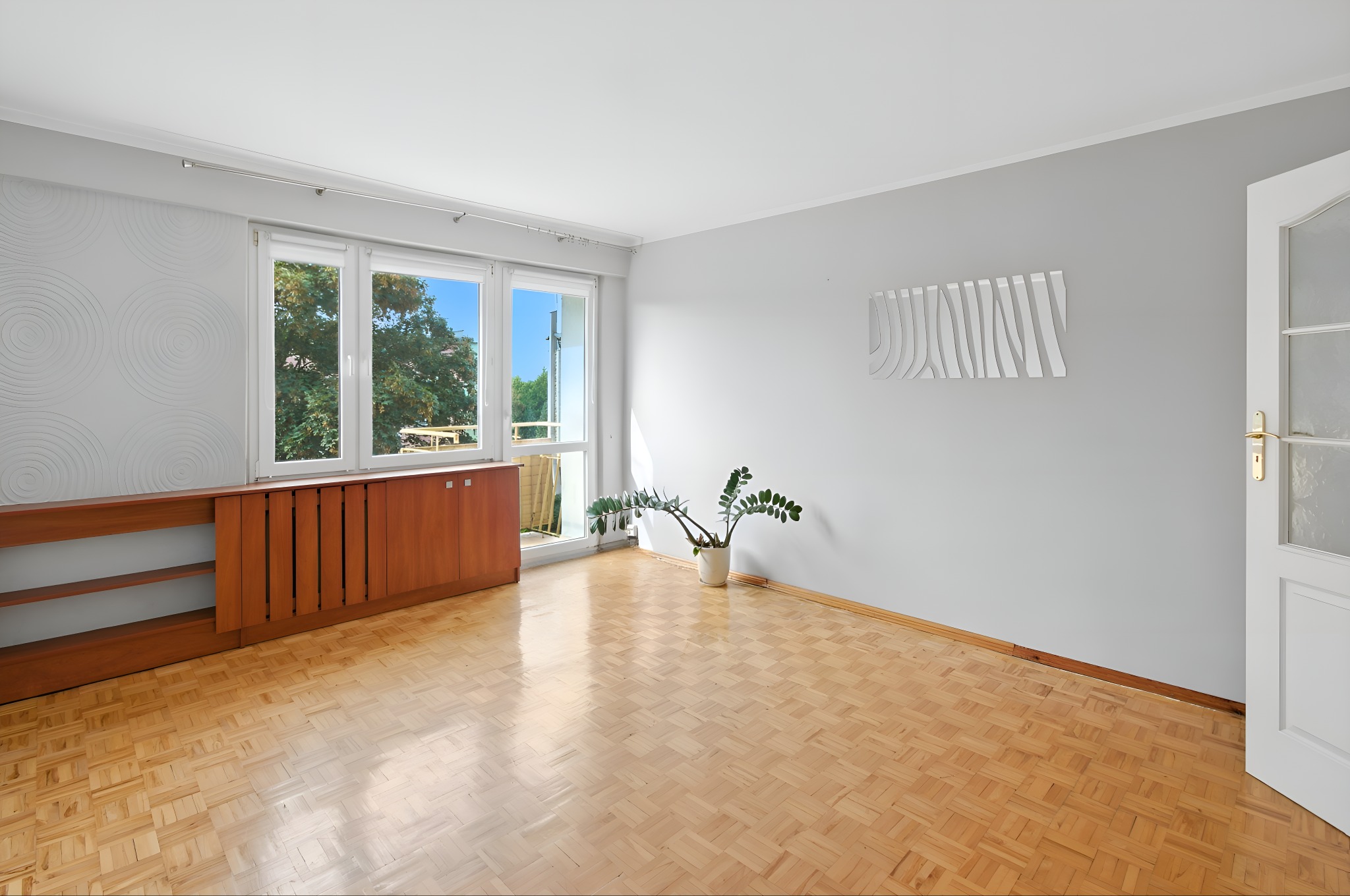 Jasne, puste wnętrze z parkietem, drewnianą szafką pod oknem i rośliną w doniczce. Widok z okna na zieleń. Białe ściany i drzwi z szybą. Minimalistyczny design.