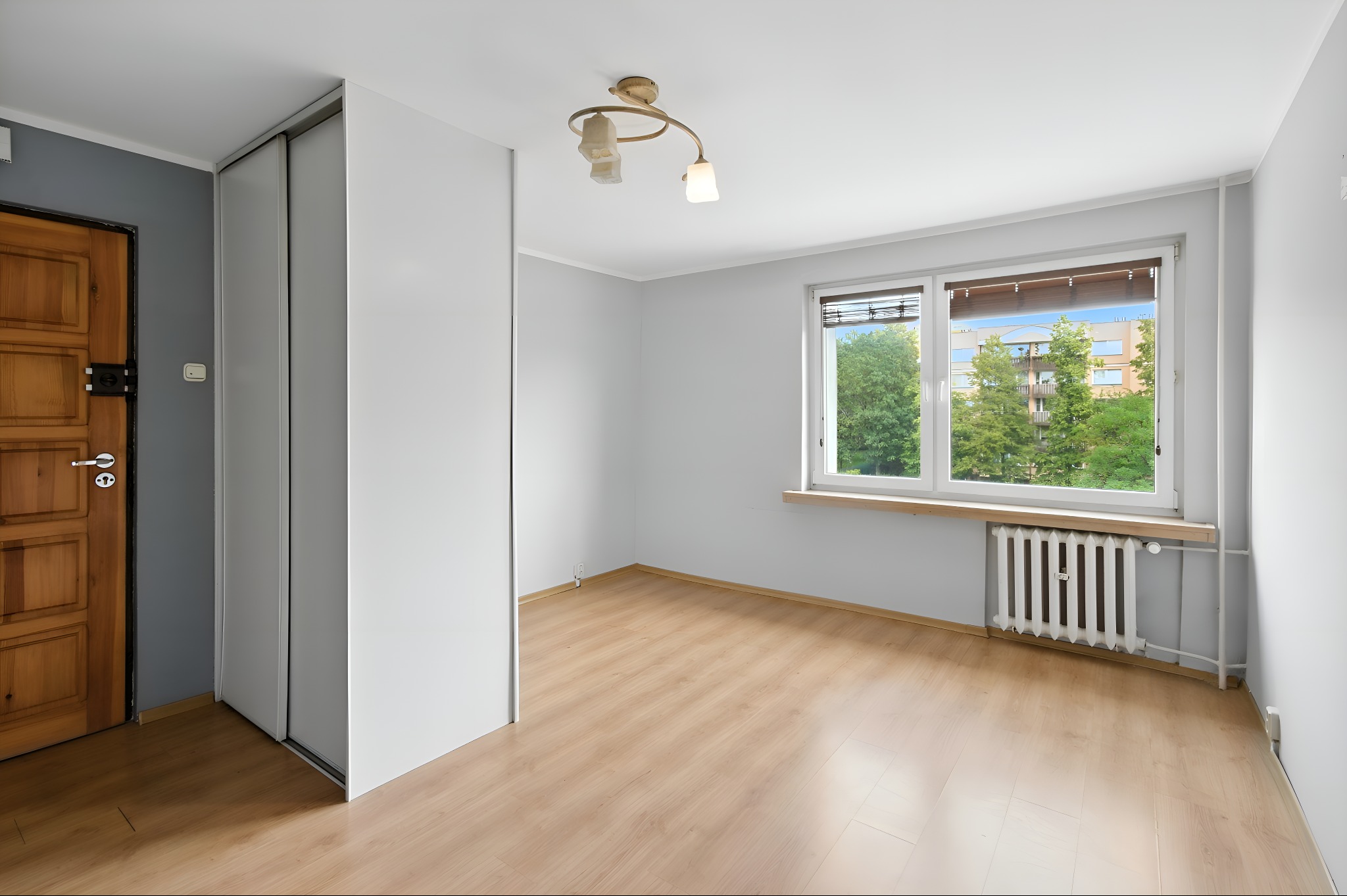 Jasne, przestronne wnętrze z drewnianą podłogą, białymi ścianami i dużym oknem z widokiem na zieleń. Widoczna szafa wnękowa i drewniane drzwi.