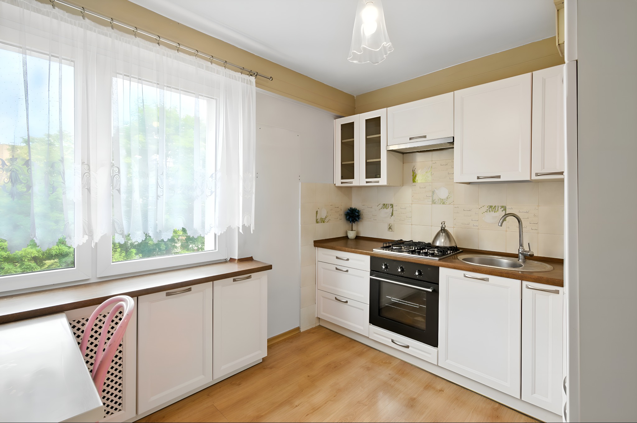 Jasna kuchnia z białymi meblami i drewnianym blatem, okno z firanką, drewniana podłoga, nowoczesny design, czarny piekarnik, srebrny zlew i bateria.