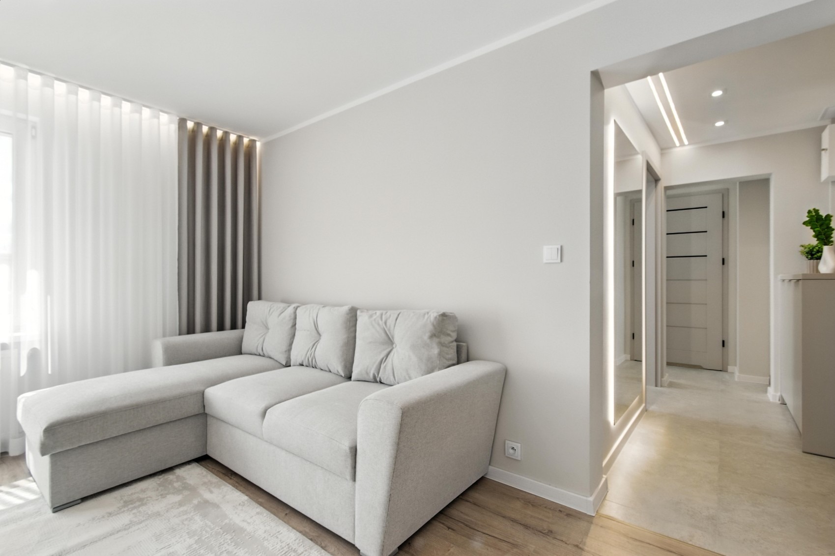 Jasne wnętrze salonu z szarą sofą narożną, zasłonami w odcieniach szarości i lustrem z podświetleniem LED. Minimalistyczny design, drewniana podłoga.