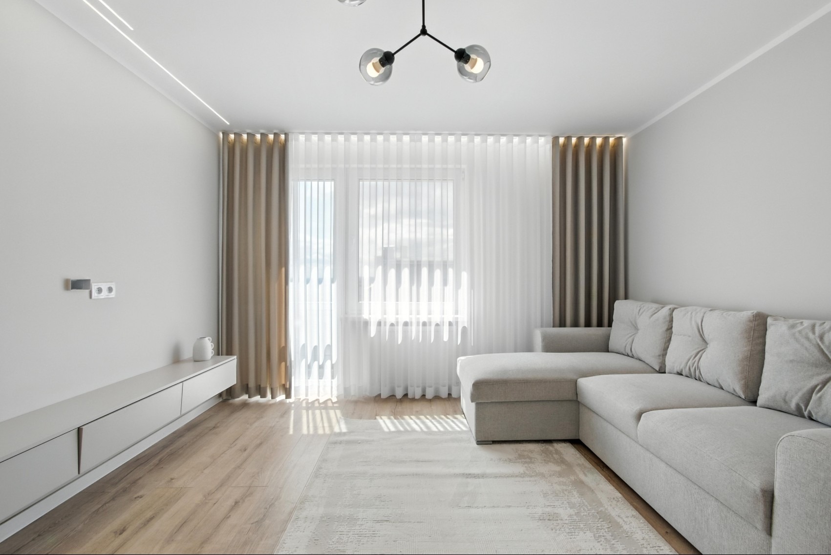 Jasne wnętrze salonu z szarą sofą narożną, drewnianą podłogą i oknem z zasłonami w odcieniach beżu i bieli. Nowoczesna lampa sufitowa.