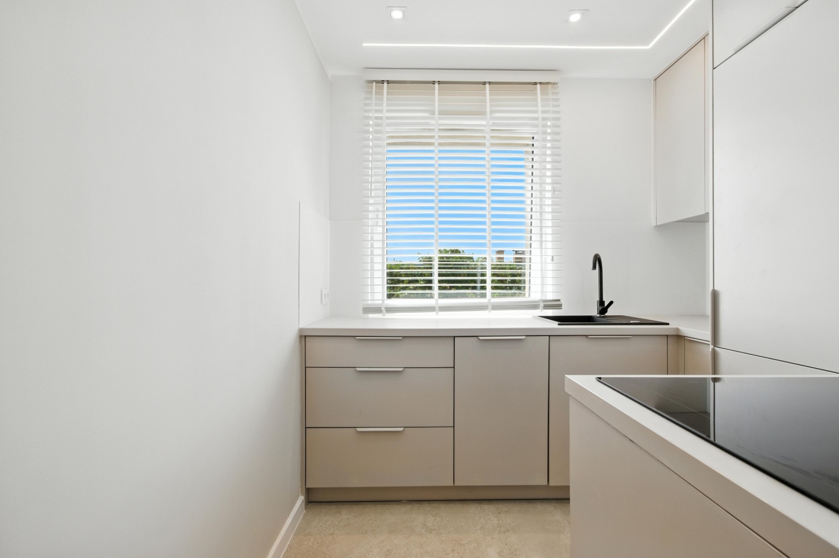 Minimalistyczna kuchnia w odcieniach bieli z akcentem czarnej armatury. Jasne blaty, białe żaluzje i widok na zieleń za oknem.