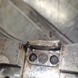 Szczegółowe ujęcie spawu na metalowej konstrukcji, z widocznymi śladami obróbki i trzema śrubami. Widoczne nierówności i detale spoiny.