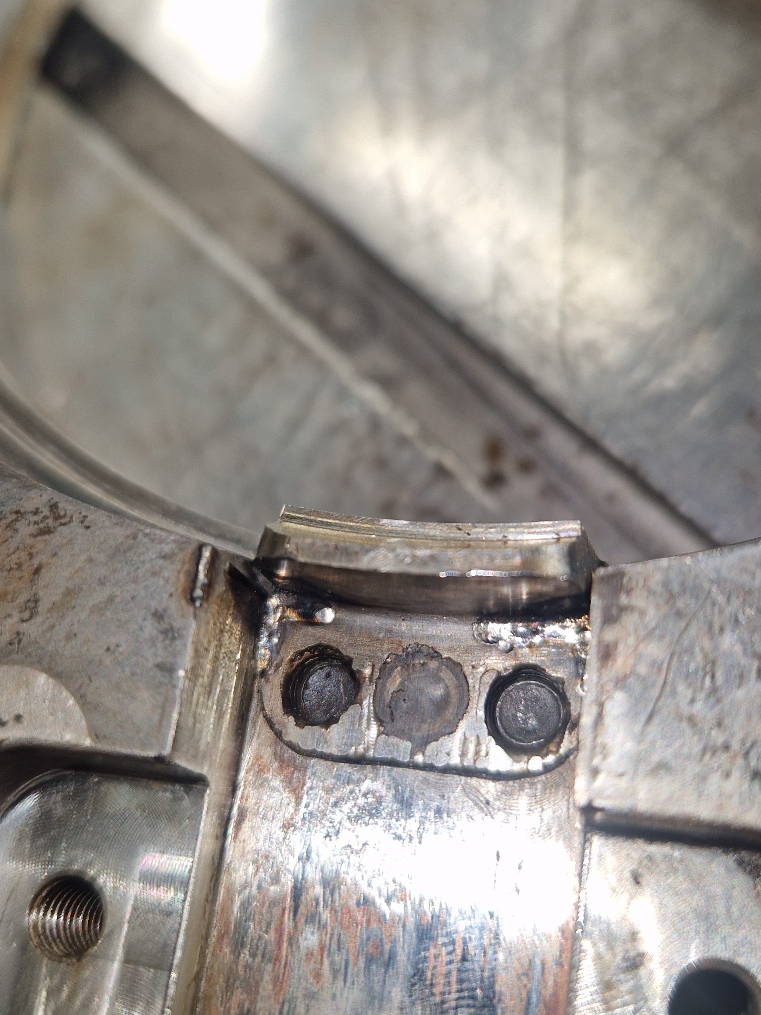 Szczegółowe ujęcie spawu na metalowej konstrukcji, z widocznymi śladami obróbki i trzema śrubami. Widoczne nierówności i detale spoiny.