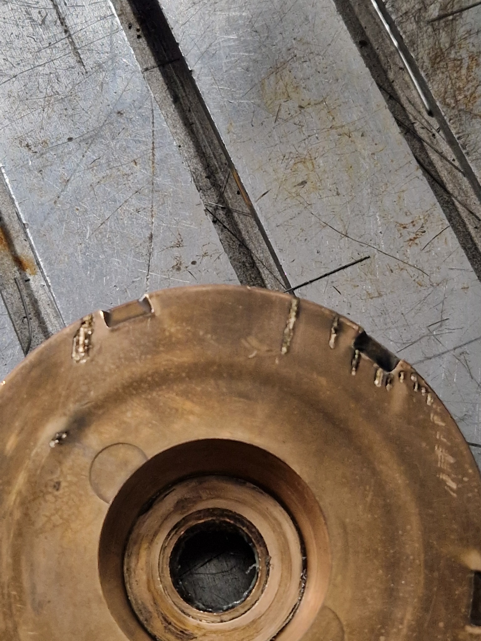 Metalowy element z wyraźnymi śladami obróbki skrawaniem i uszkodzeniami na krawędziach, leżący na metalowym stole warsztatowym z widocznymi zarysowaniami.