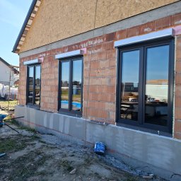 OKNA DRZWI FASADY YURII ROZHKO - Nowe, grafitowe okna z roletami zewnętrznymi w surowym stanie budynku z cegły. Widoczne detale montażu i fragment terenu budowy.