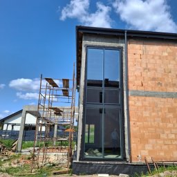 OKNA DRZWI FASADY YURII ROZHKO - Wysokie okno z czarną ramą w surowym budynku z cegły, widoczne rusztowanie i błękitne niebo. Okno odbija otoczenie, w tym postać wewnątrz.