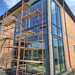 OKNA DRZWI FASADY YURII ROZHKO - Montaż dużych, panoramicznych okien w surowym budynku z cegły. Widoczne rusztowania i czarne ramy okienne. Nowoczesna architektura w trakcie realizacji.
