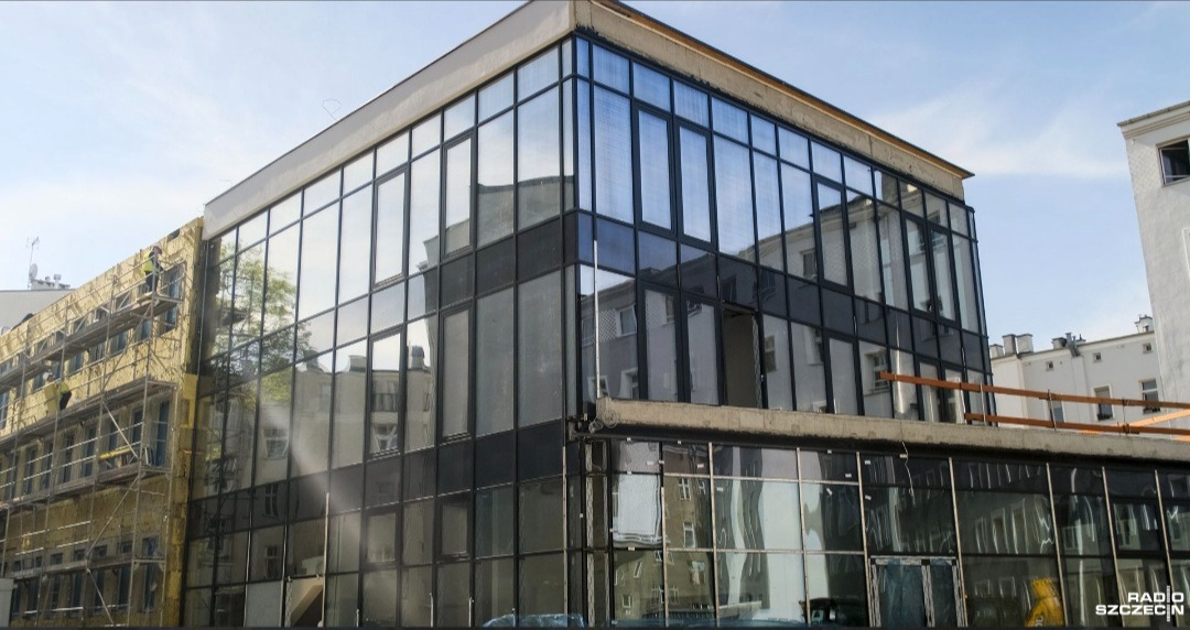 Budynek z przeszkloną fasadą w trakcie prac wykończeniowych. Część budynku w rusztowaniach, widoczne okna i odbicia innych budynków w szklanej elewacji.