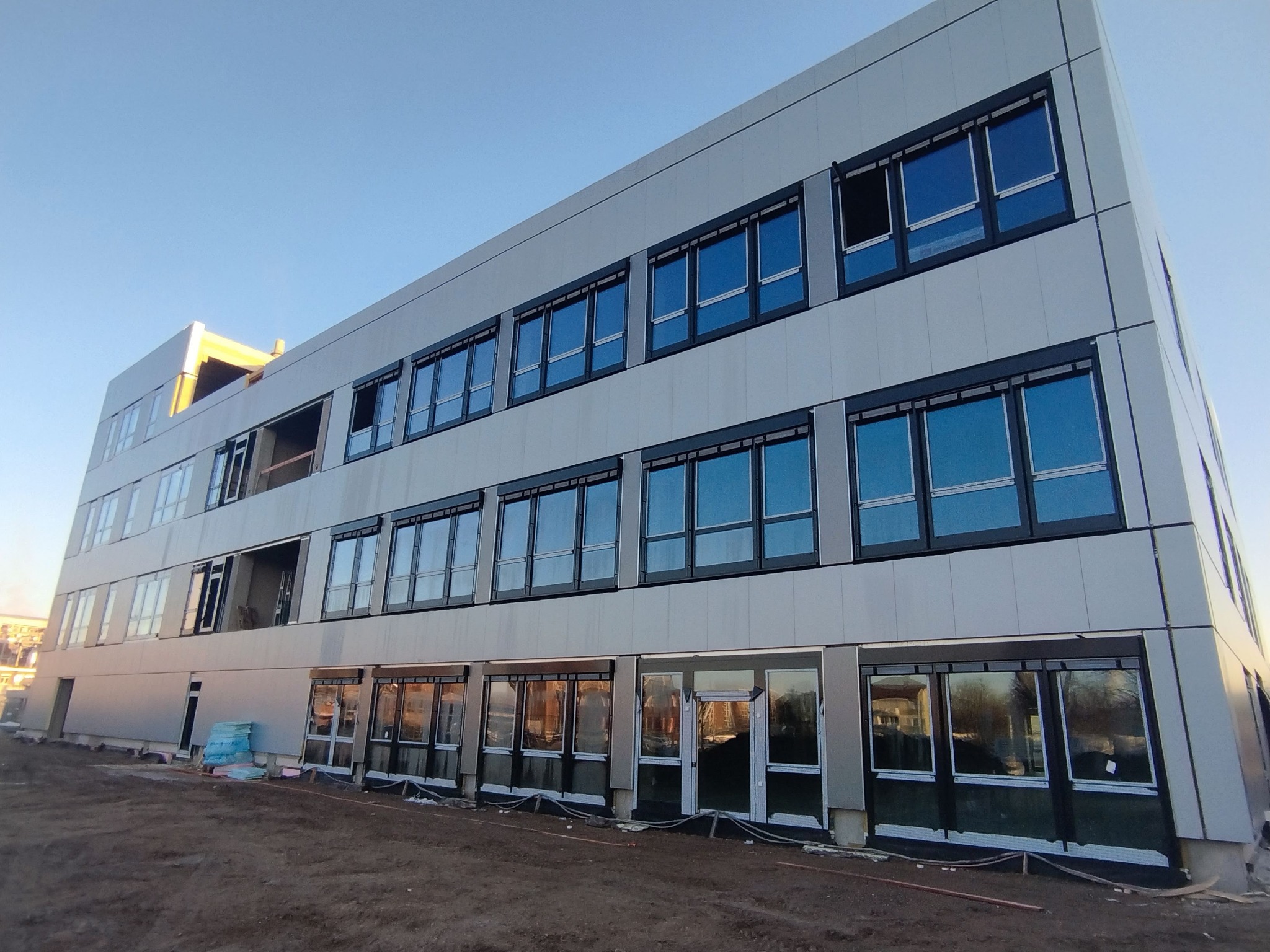 Nowoczesny budynek z zamontowanymi oknami i drzwiami, widoczne prace wykończeniowe na zewnątrz, błękitne niebo w tle. Część okien jeszcze nieotwarta.