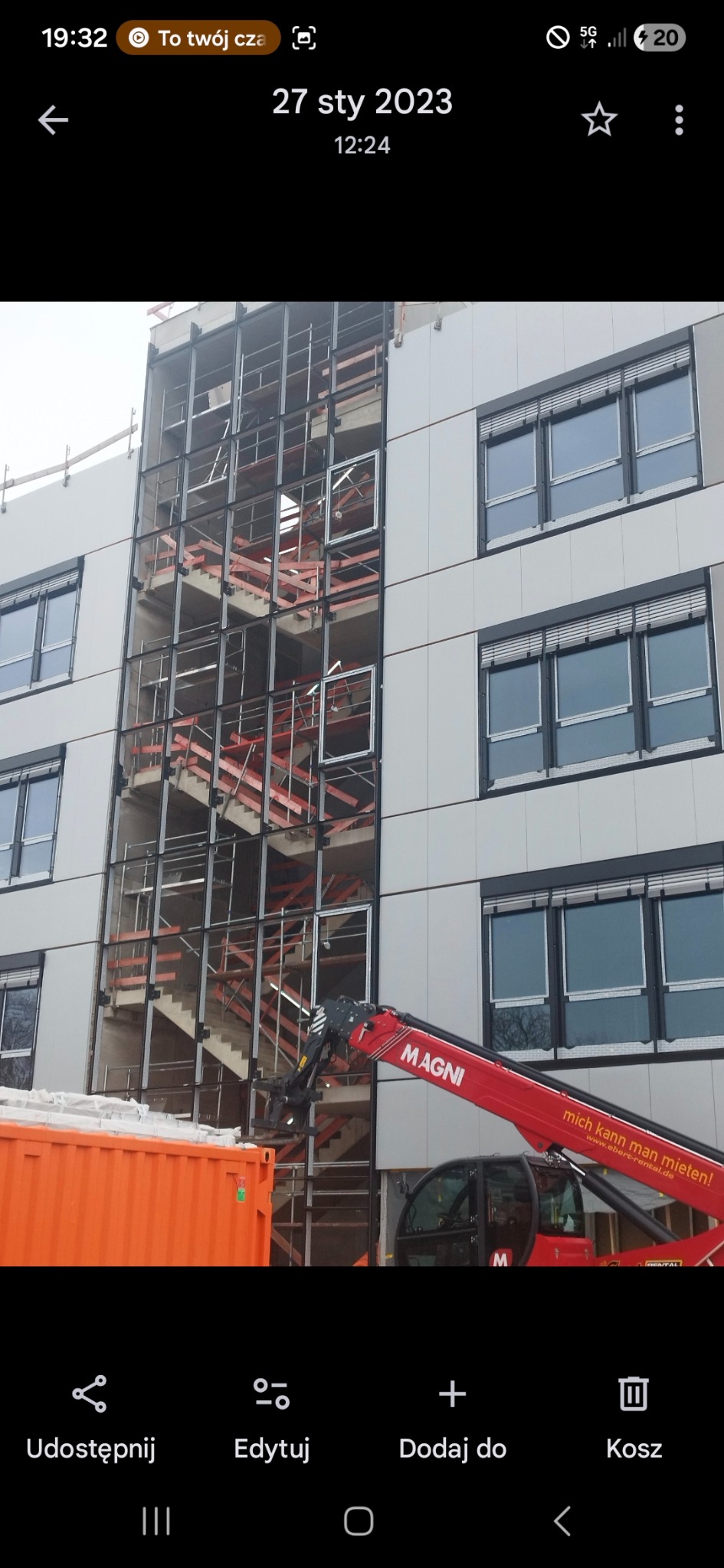 Budynek w trakcie montażu okien i konstrukcji schodów zewnętrznych. Widoczny dźwig MAGNI i kontener. Szare panele elewacyjne i częściowo otwarte okna.