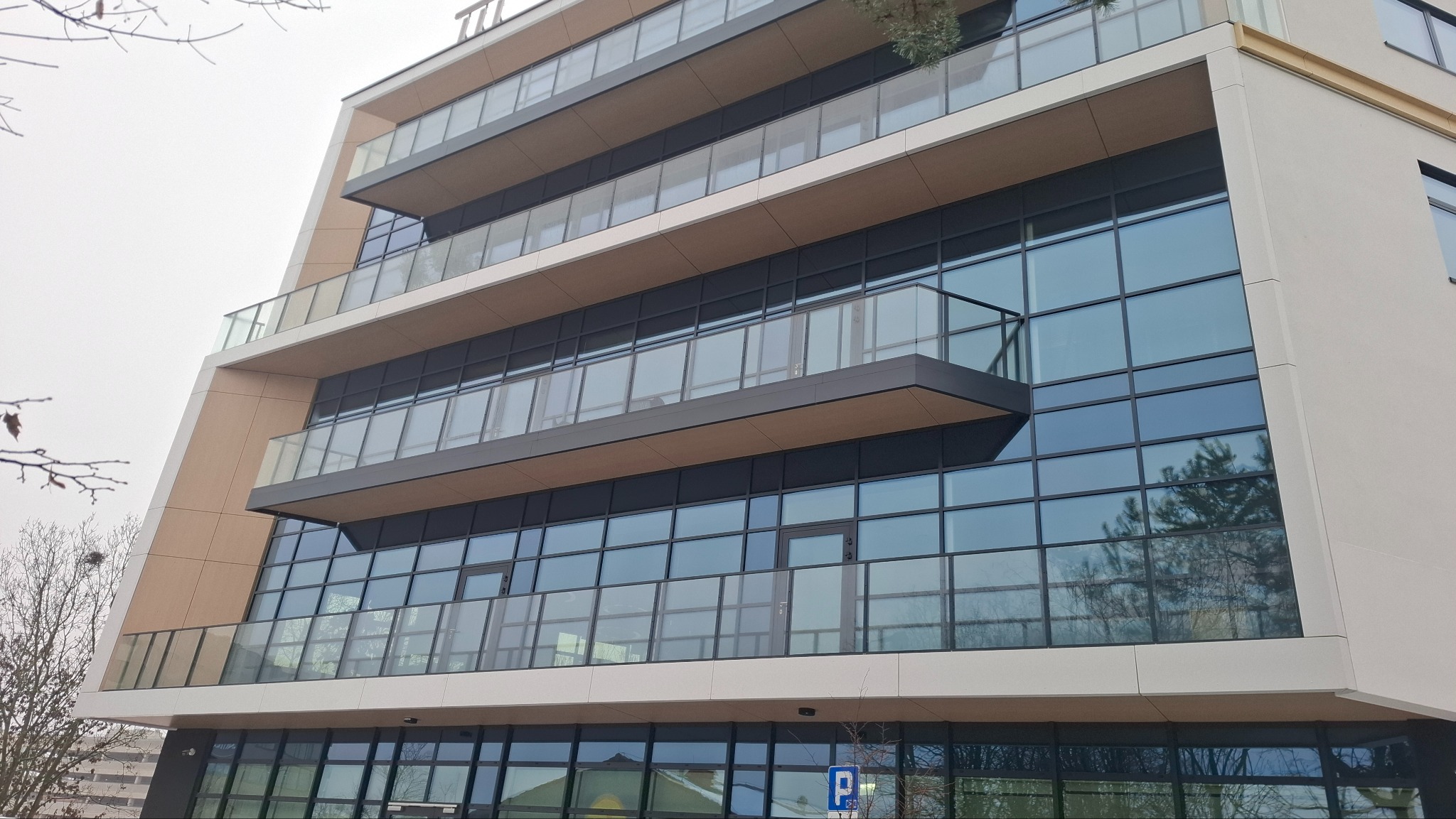 Nowoczesny budynek z balkonami i dużymi oknami, widok z dołu, szklane balustrady, minimalistyczna architektura, pochmurne niebo, drzewa bez liści w tle.