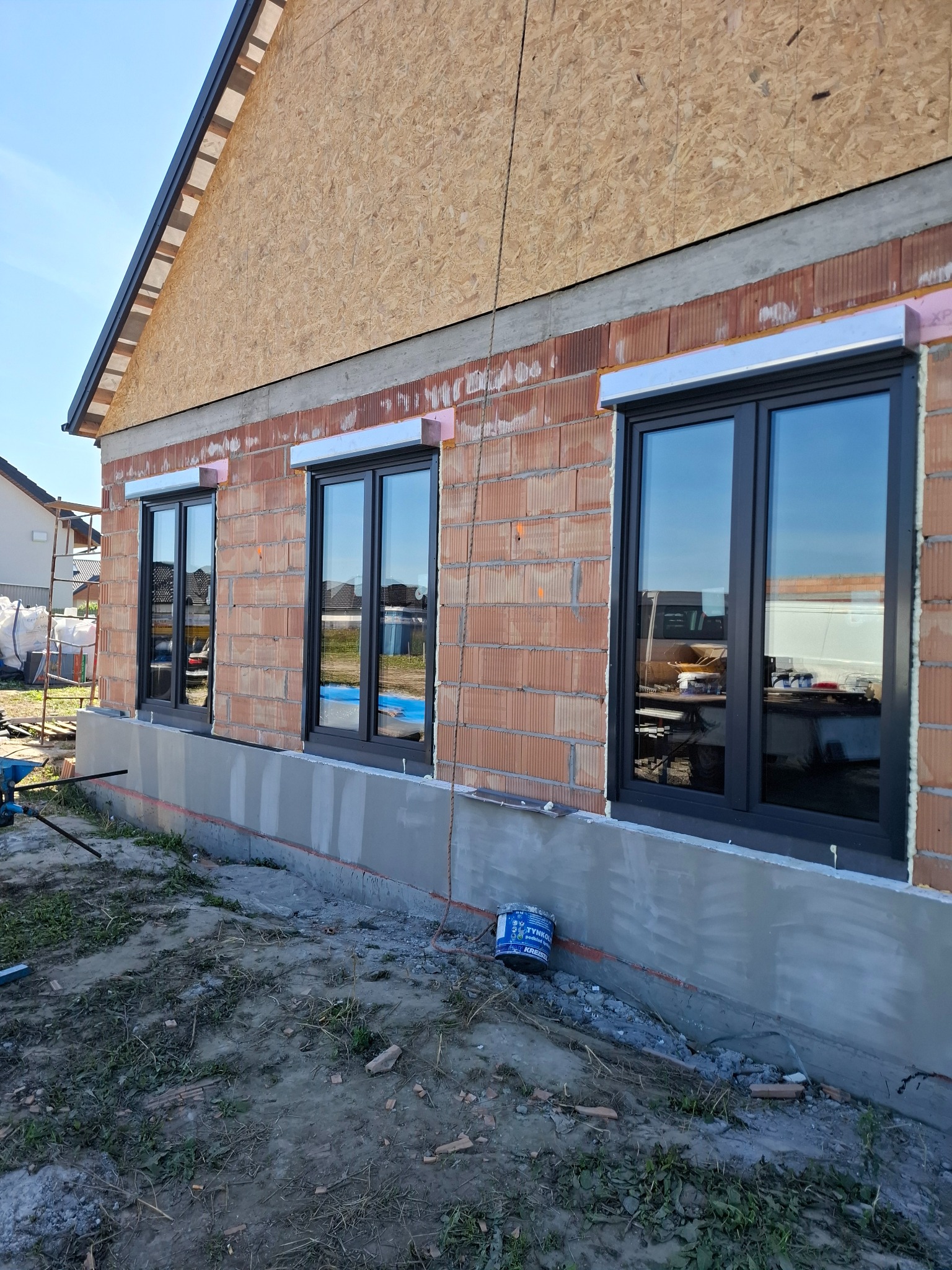 Nowe, grafitowe okna z roletami zewnętrznymi w surowym stanie budynku z cegły. Widoczne detale montażu i fragment terenu budowy.
