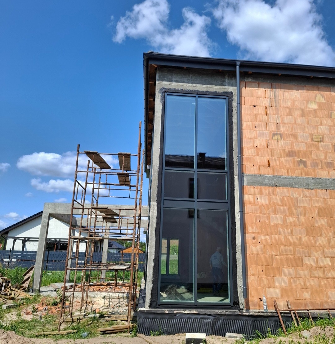 Wysokie okno z czarną ramą w surowym budynku z cegły, widoczne rusztowanie i błękitne niebo. Okno odbija otoczenie, w tym postać wewnątrz.