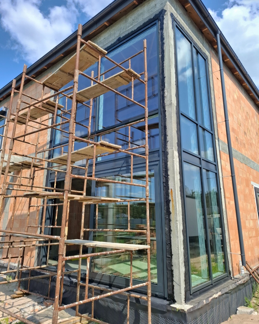 Montaż dużych, panoramicznych okien w surowym budynku z cegły. Widoczne rusztowania i czarne ramy okienne. Nowoczesna architektura w trakcie realizacji.