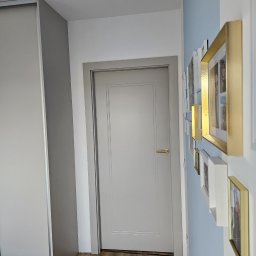 Radosław Kostka - Szara, klasyczna wewnętrzna stolarka drzwiowa w mieszkaniu. Na ścianie galeria ramek. Podłoga z paneli imitujących drewno. Minimalistyczny design.