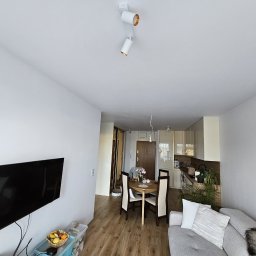 Radosław Kostka - Jasne wnętrze mieszkania po remoncie: ściany pomalowane na biało, drewniana podłoga, nowoczesne oświetlenie sufitowe, telewizor na ścianie i widoczny fragment sofy.