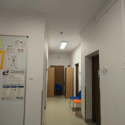Radosław Kostka - Korytarz w placówce medycznej z białymi ścianami, linoleum na podłodze, oświetleniem sufitowym i meblami w odcieniach brązu i niebieskiego. Na ścianie tablica informacyjna.