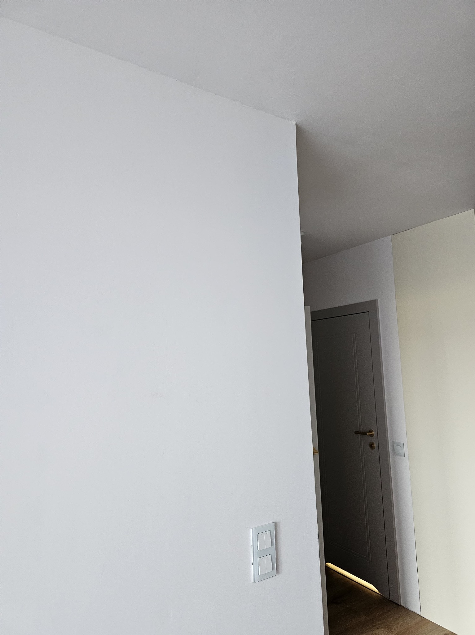 Ściana po malowaniu na biało z widocznym włącznikiem światła. W tle drzwi i fragment podłogi. Minimalistyczne wykończenie wnętrza.