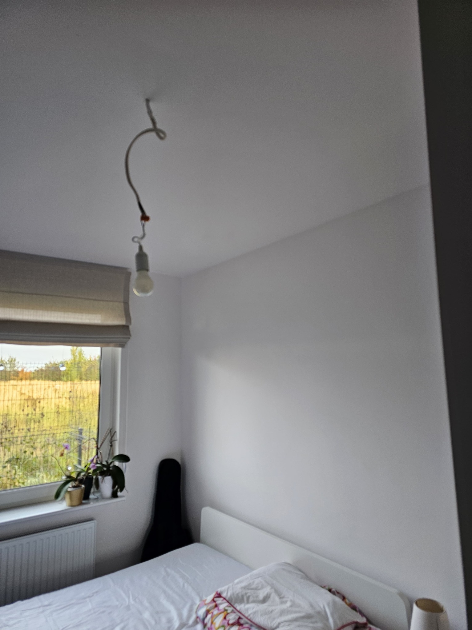 Pokój po malowaniu ścian na biało, widoczna żarówka na kablu, okno z roletą rzymską, kwiaty na parapecie, futerał na gitarę i łóżko z białą pościelą.