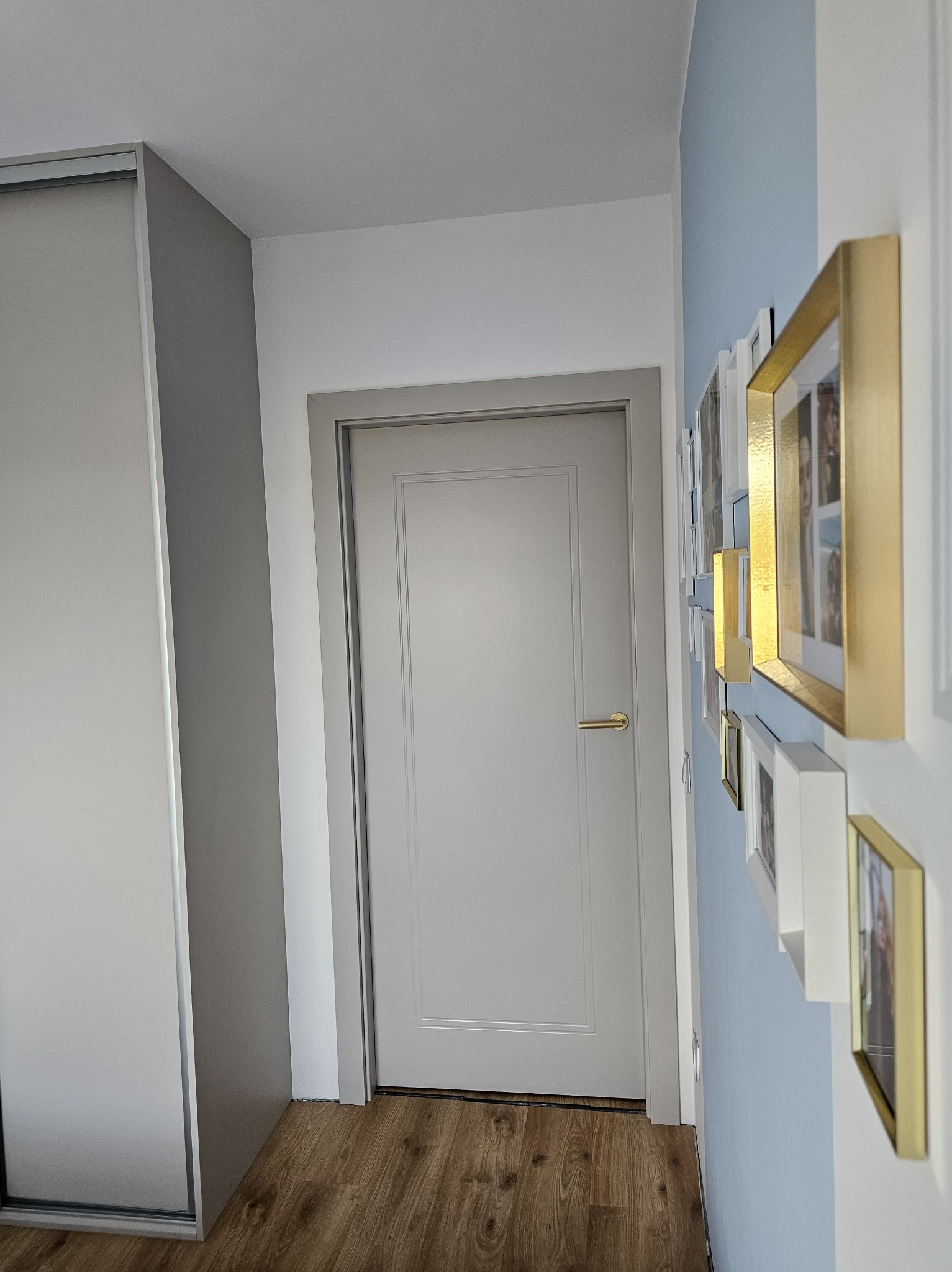 Szara, klasyczna wewnętrzna stolarka drzwiowa w mieszkaniu. Na ścianie galeria ramek. Podłoga z paneli imitujących drewno. Minimalistyczny design.