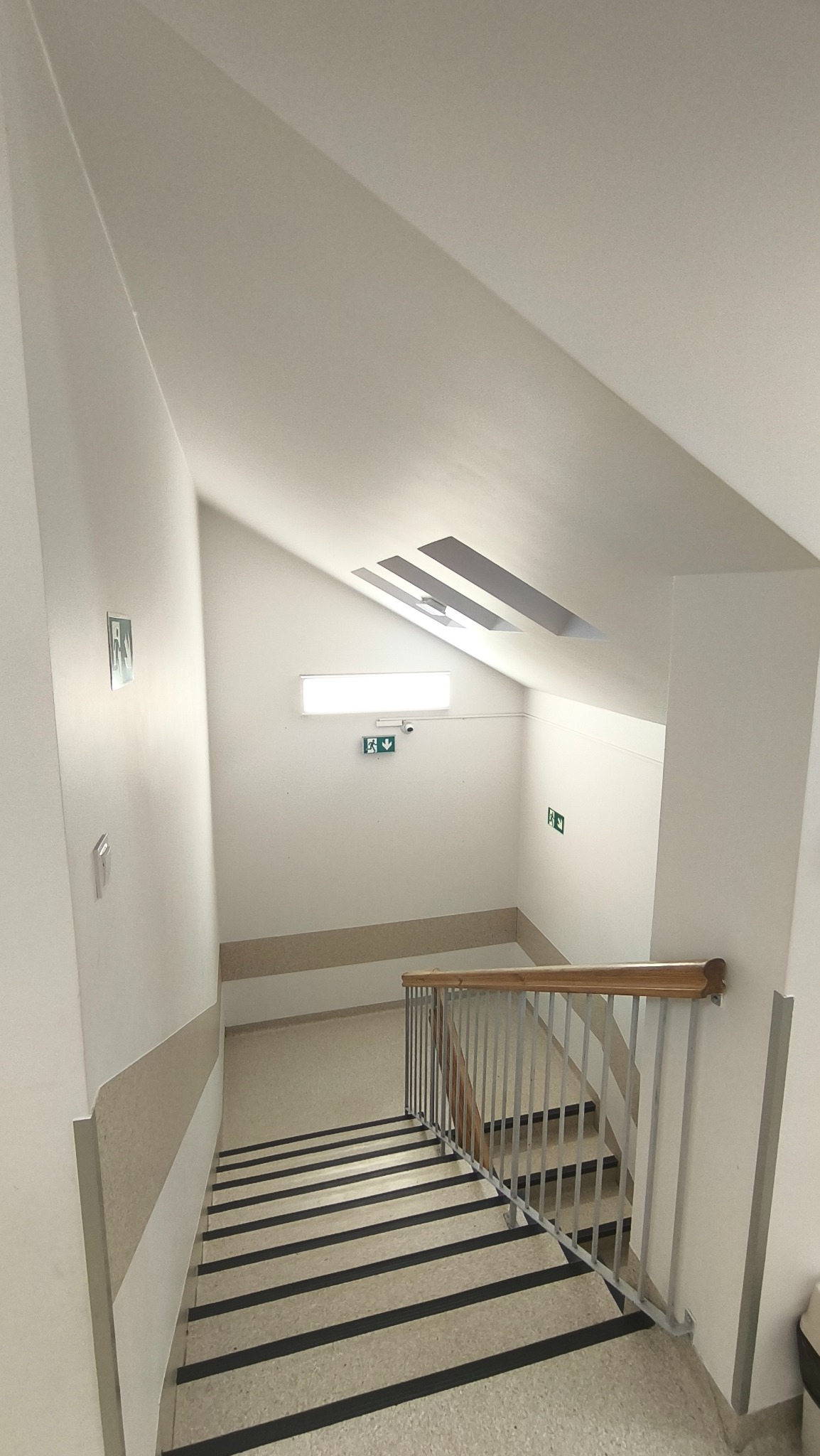 Jasne schody z drewnianą poręczą i metalowymi prętami, widok z góry. Białe ściany z oznaczeniami ewakuacyjnymi i beżową lamperią. Oświetlenie sufitowe.