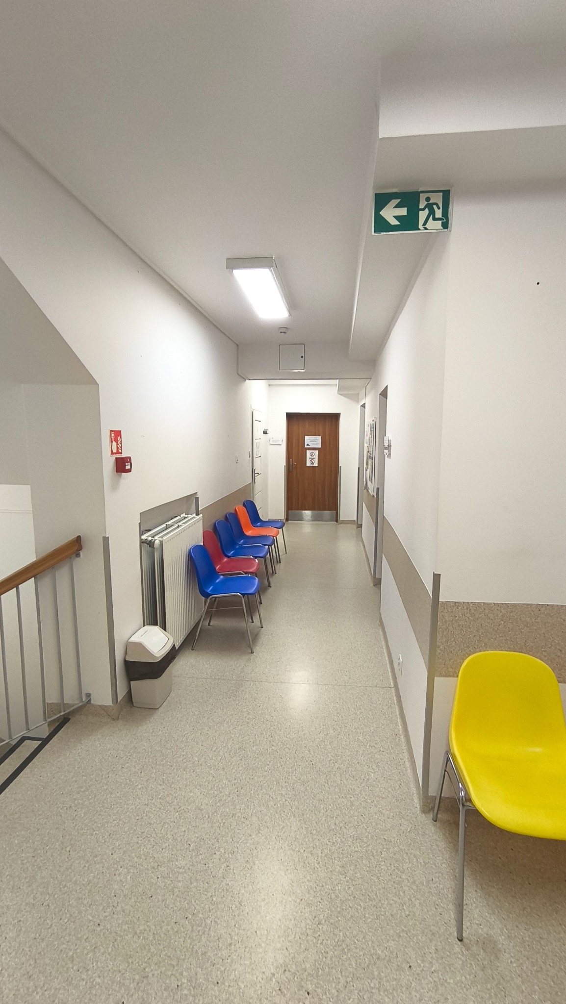 Korytarz z krzesłami w kolorach niebieskim, pomarańczowym i żółtym. Na ścianie znak ewakuacyjny i gaśnica. Podłoga z lastryko. Oświetlenie sufitowe.