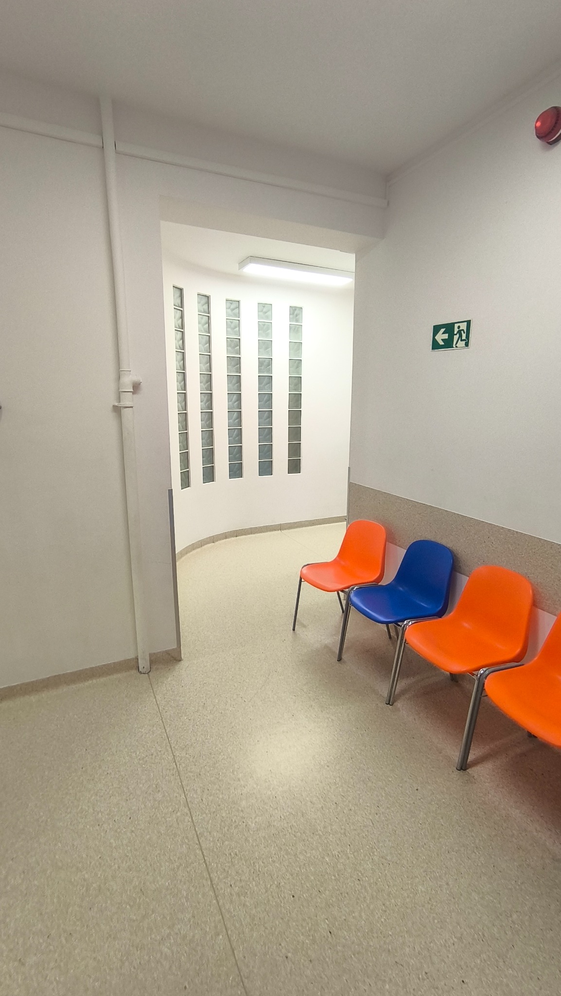 Korytarz z trzema krzesłami (pomarańczowe i niebieskie) ustawionymi pod ścianą. Na ścianie znak ewakuacyjny i lampa. Podłoga wyłożona jasnym linoleum.