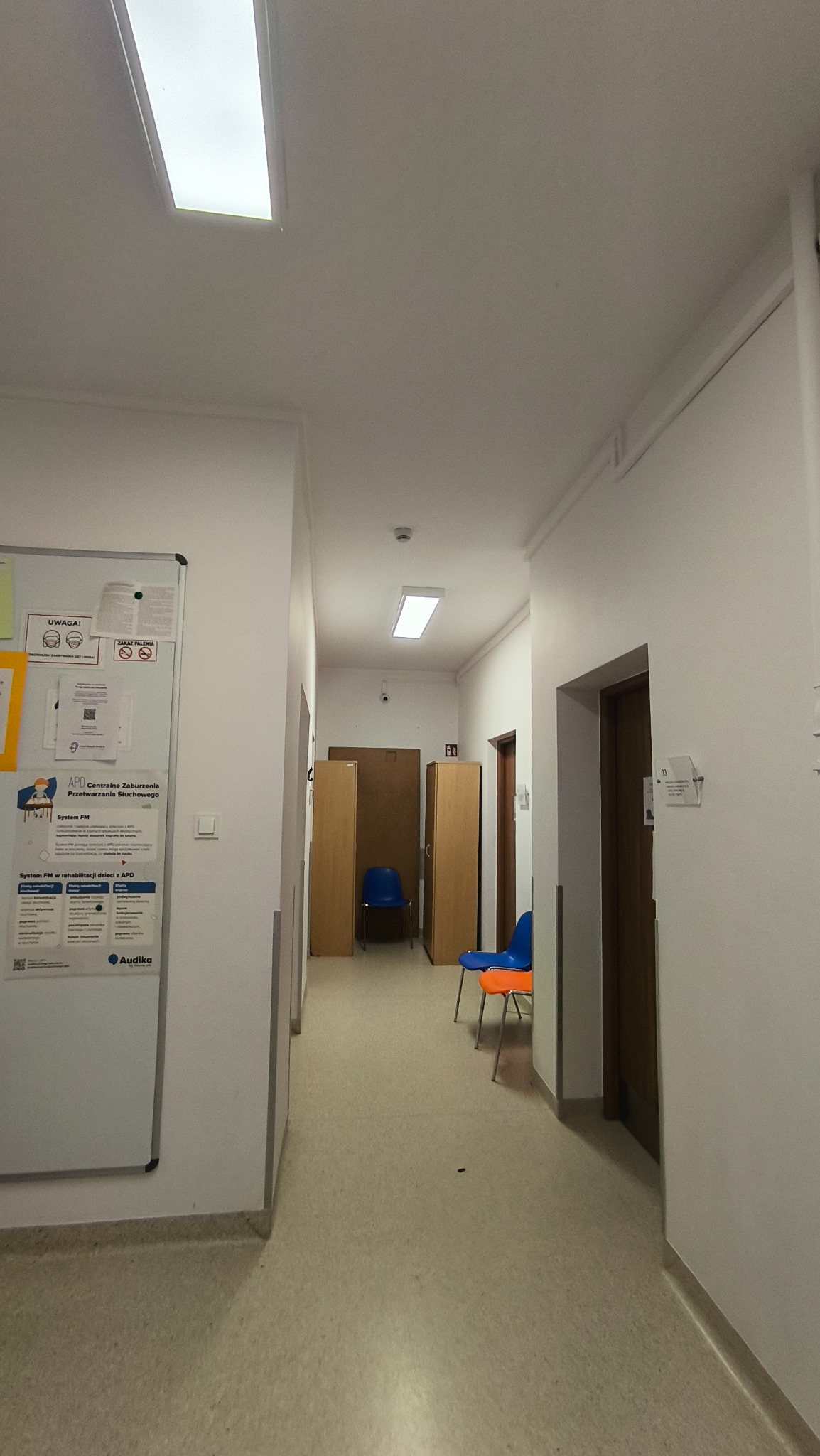 Korytarz w placówce medycznej z białymi ścianami, linoleum na podłodze, oświetleniem sufitowym i meblami w odcieniach brązu i niebieskiego. Na ścianie tablica informacyjna.
