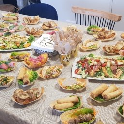 Gastrochef - Stół zastawiony różnorodnymi daniami cateringowymi w drewnianych miseczkach i na talerzach, gotowy na przyjęcie urodzinowe. Widoczne sałatki, spring rollsy, tacos i przekąski.