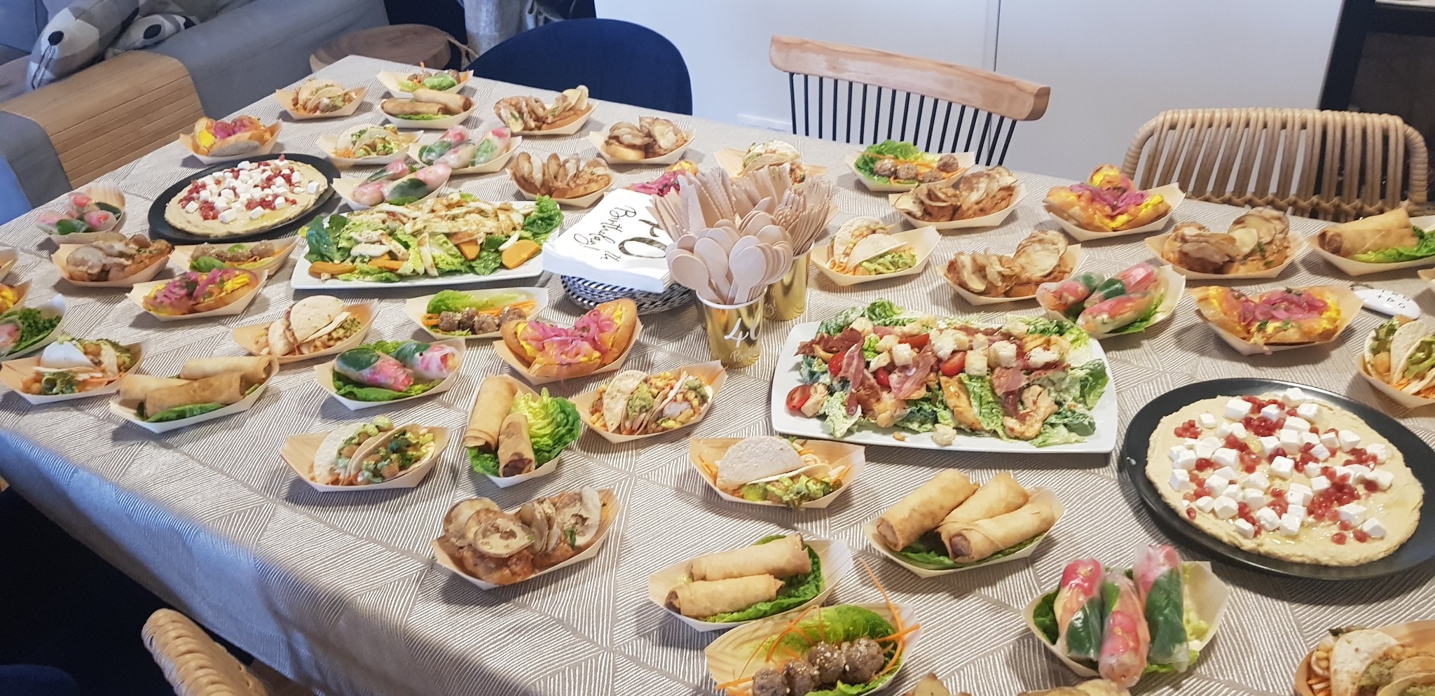 Stół zastawiony różnorodnymi daniami cateringowymi w drewnianych miseczkach i na talerzach, gotowy na przyjęcie urodzinowe. Widoczne sałatki, spring rollsy, tacos i przekąski.