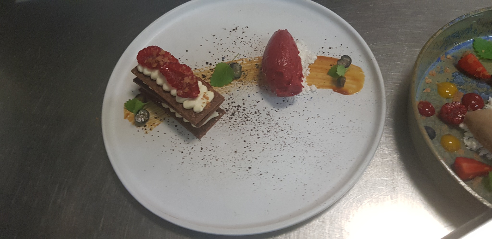 Wykwintny deser na białym talerzu: warstwowy przekładaniec z kremem i malinami obok sorbetu malinowego, udekorowany sosem i posypką. Elegancka kompozycja.