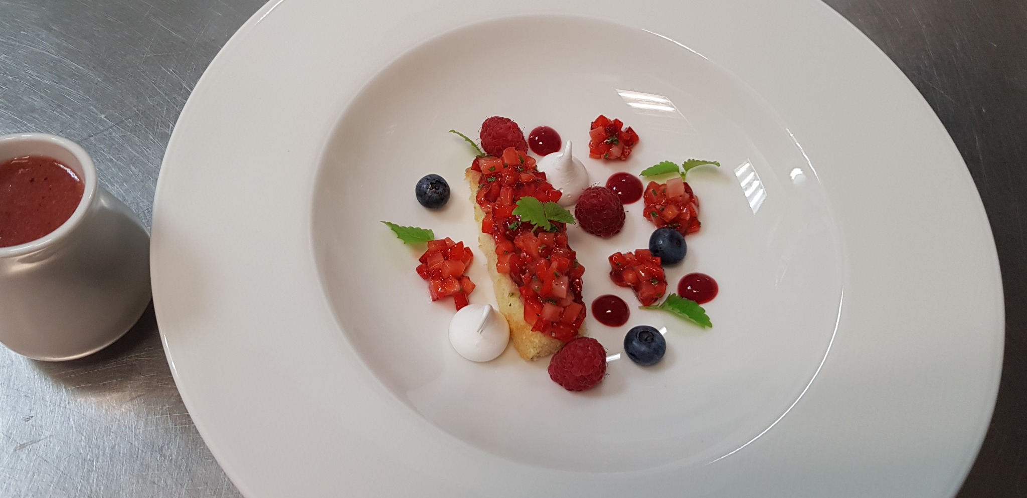 Elegancko podane danie deserowe na białym talerzu: tost z truskawkami, maliny, borówki, bezy i sos. Fine dining, nowoczesna prezentacja, metalowe tło.