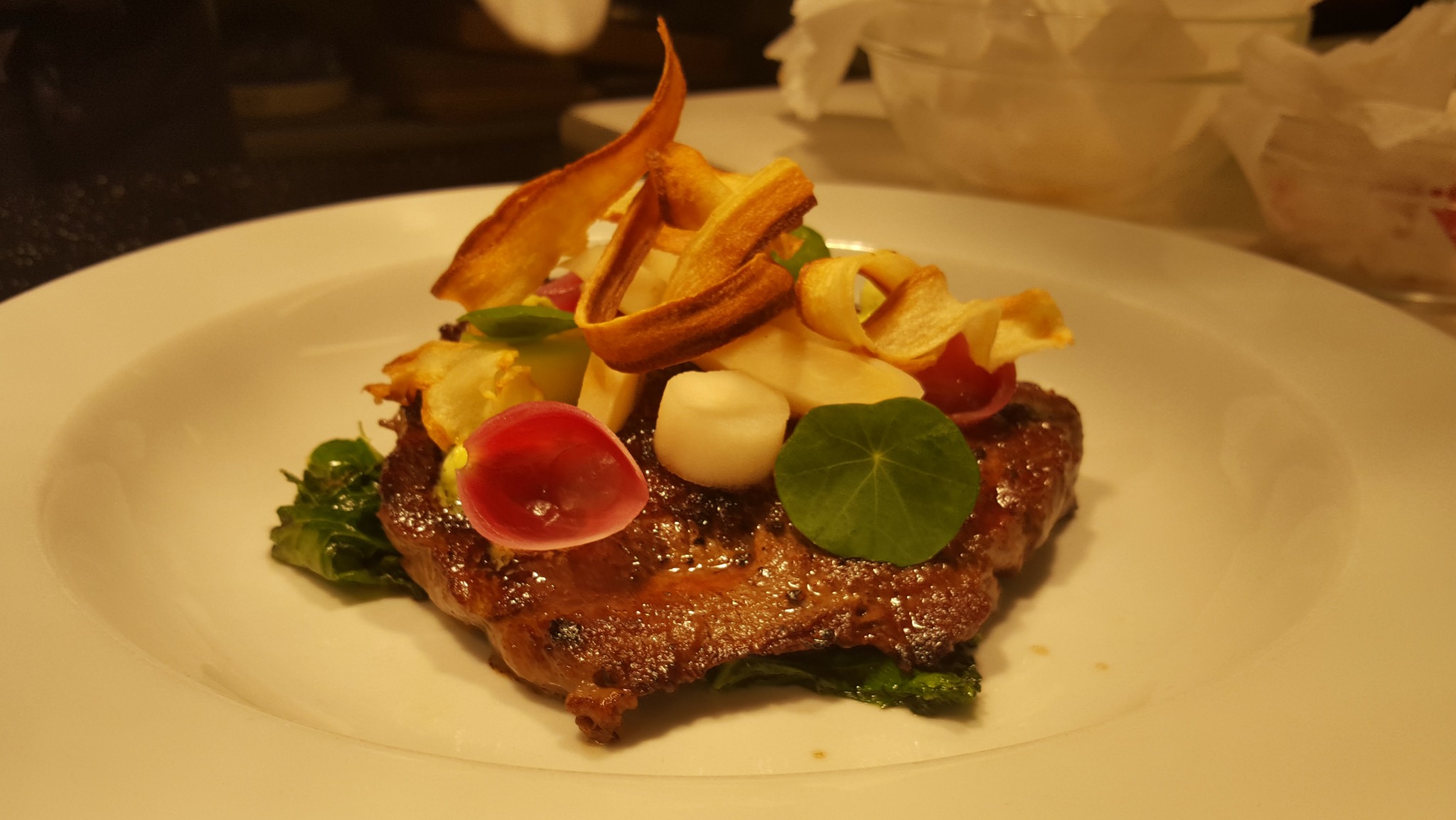 Wykwintne danie mięsne na białym talerzu, udekorowane warzywami, chipsami i ziołami. Elegancki sposób podania, światło podkreśla detale tekstury potrawy.