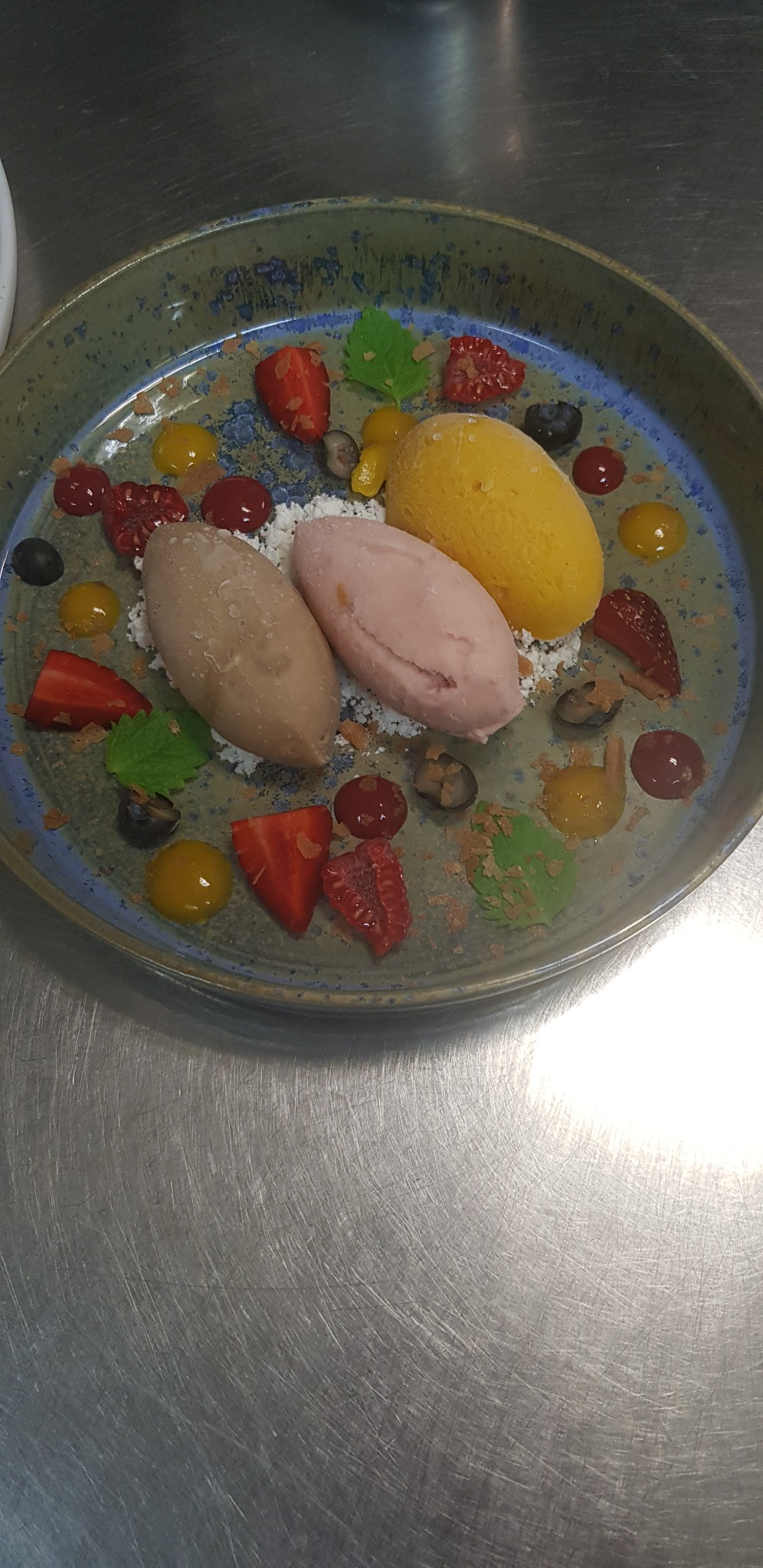 Elegancko podane lody w trzech smakach na talerzu z owocami i sosem. Lody o smaku czekoladowym, truskawkowym i mango. Dekoracja z malin, truskawek i borówek.