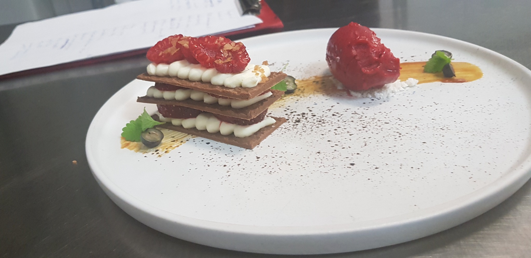 Elegancki deser: warstwowy torcik z malinami i kremem obok sorbetu na białym talerzu, z dekoracją z borówek i mięty, na metalowym blacie.