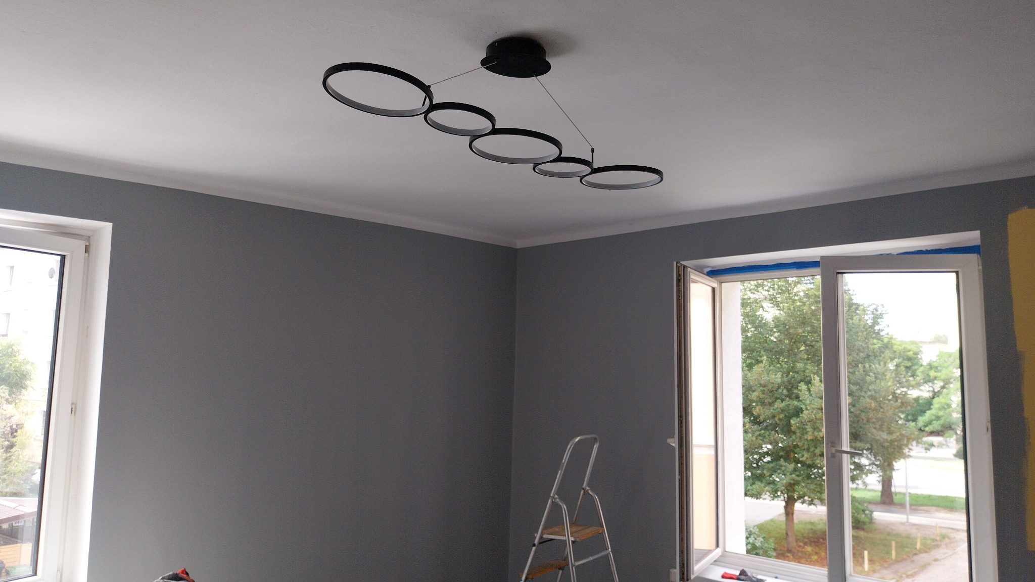 Szare ściany po malowaniu w pokoju z nowoczesną lampą pierścieniową. Drabina stoi obok okna z widokiem na zieleń. Wykończenie wnętrza w trakcie.