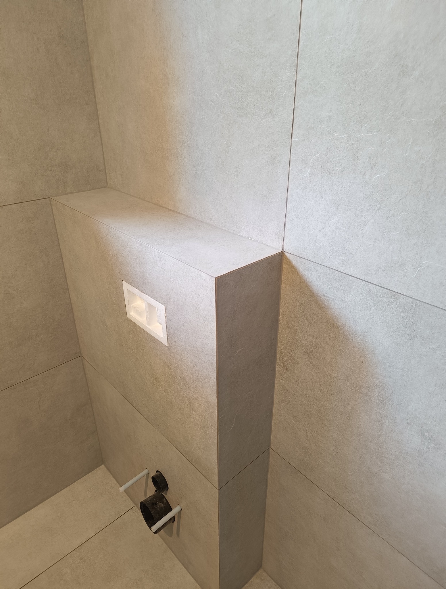 Wykończona łazienka: szare, matowe płytki, zabudowa stelaża WC z wbudowanym przyciskiem, widoczne przyłącza kanalizacyjne. Minimalistyczny design.