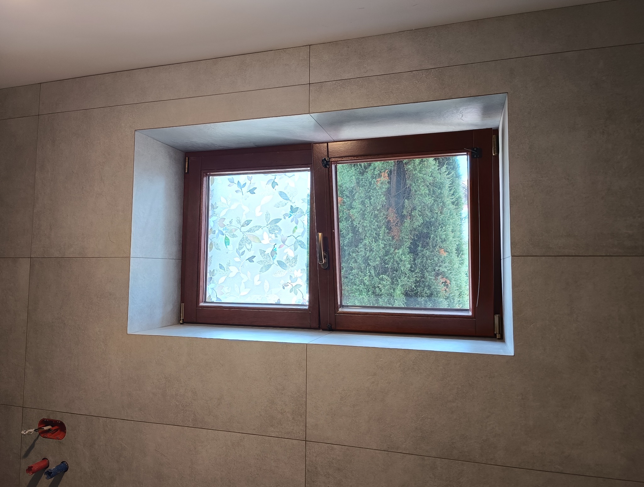 Wykończona łazienka z oknem w brązowej ramie. Na szybie dekoracyjna folia z motywem liści. Beżowe, matowe płytki na ścianach. Widoczne przyłącza wodne.