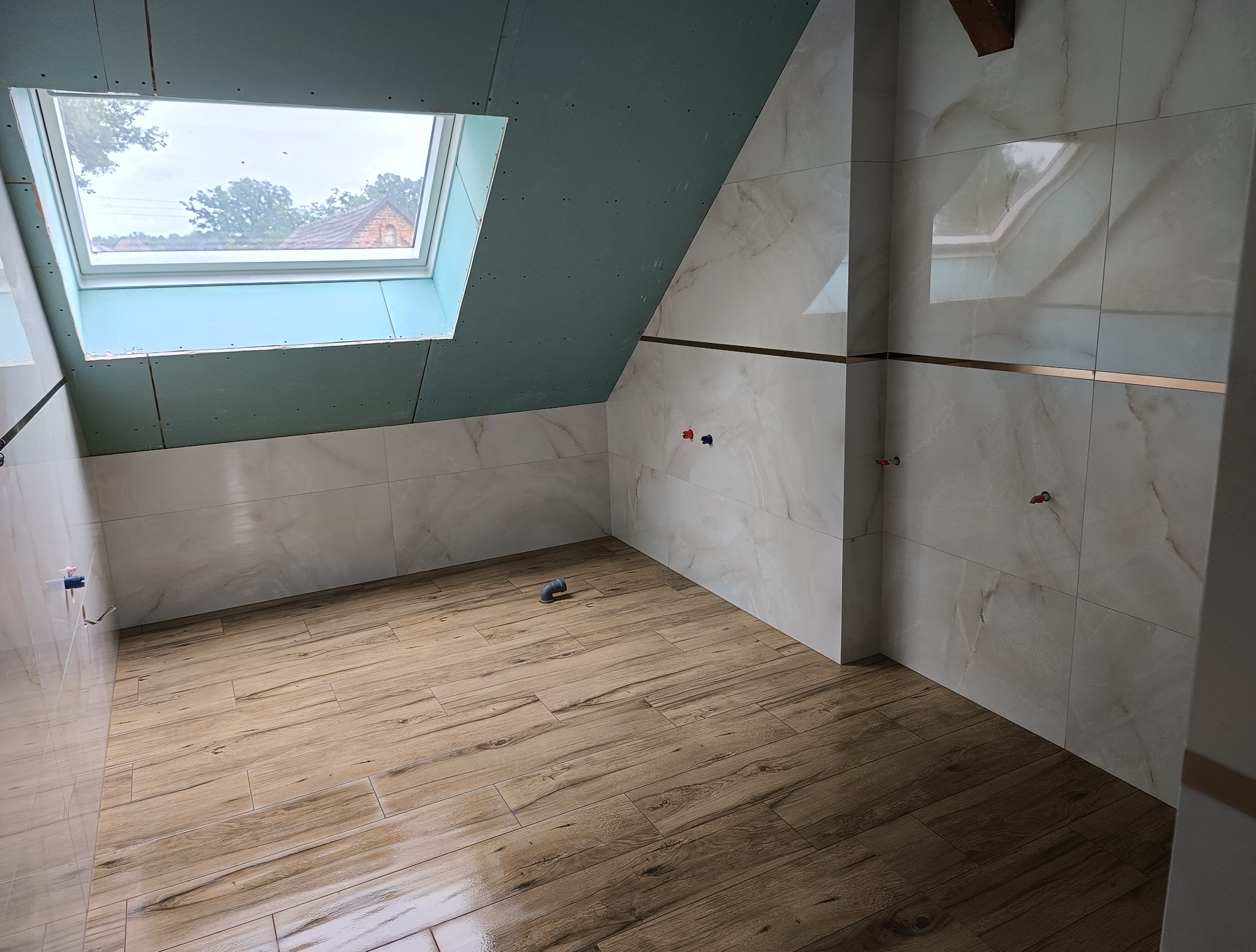 Wykończona łazienka na poddaszu z oknem dachowym i płytkami imitującymi drewno na podłodze oraz marmur na ścianach, z dekoracyjnymi listwami.