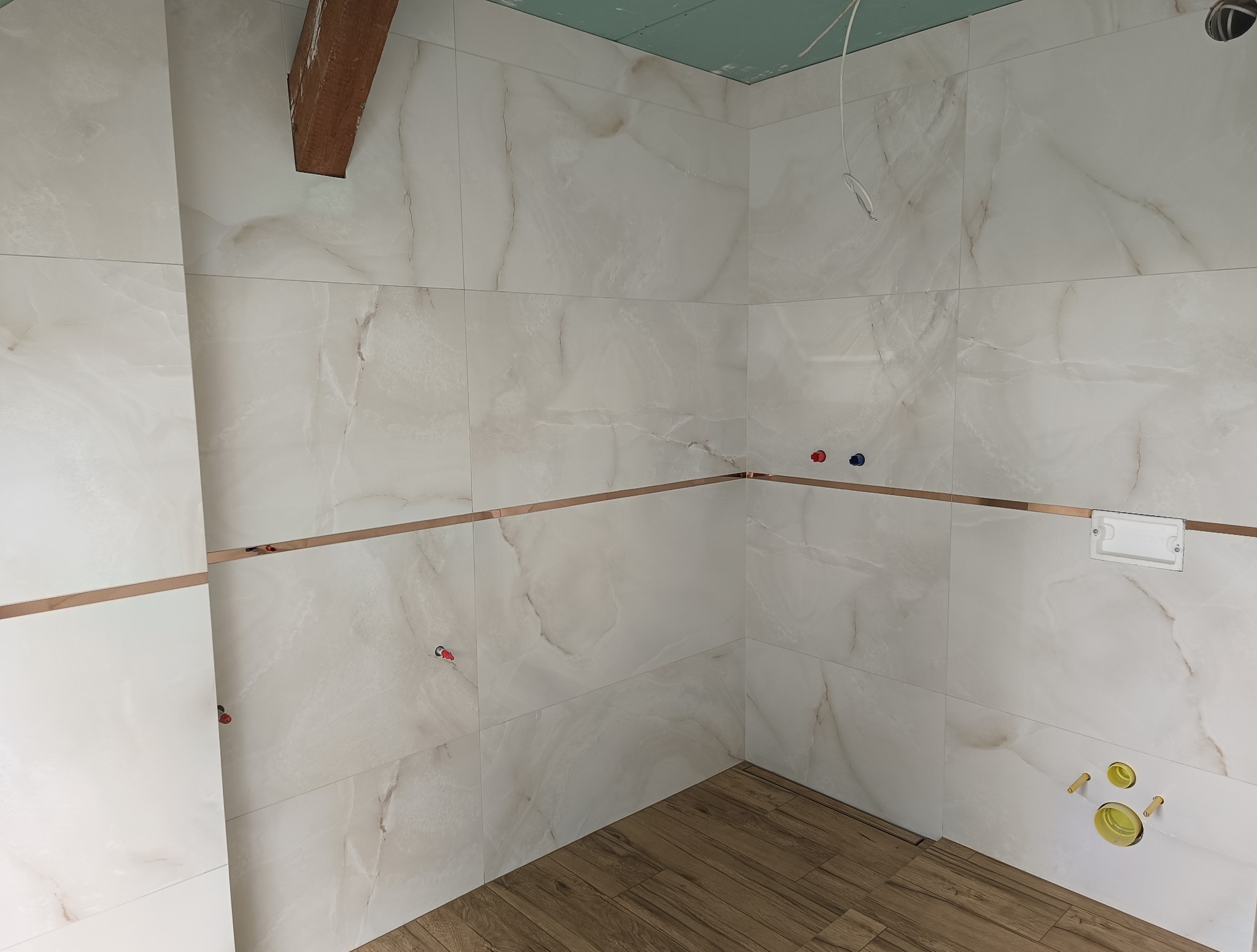 Wykończona łazienka z marmurowymi płytkami, akcentem w postaci ozdobnej listwy i drewnopodobną podłogą. Widoczne przyłącza i stelaż podtynkowy WC.